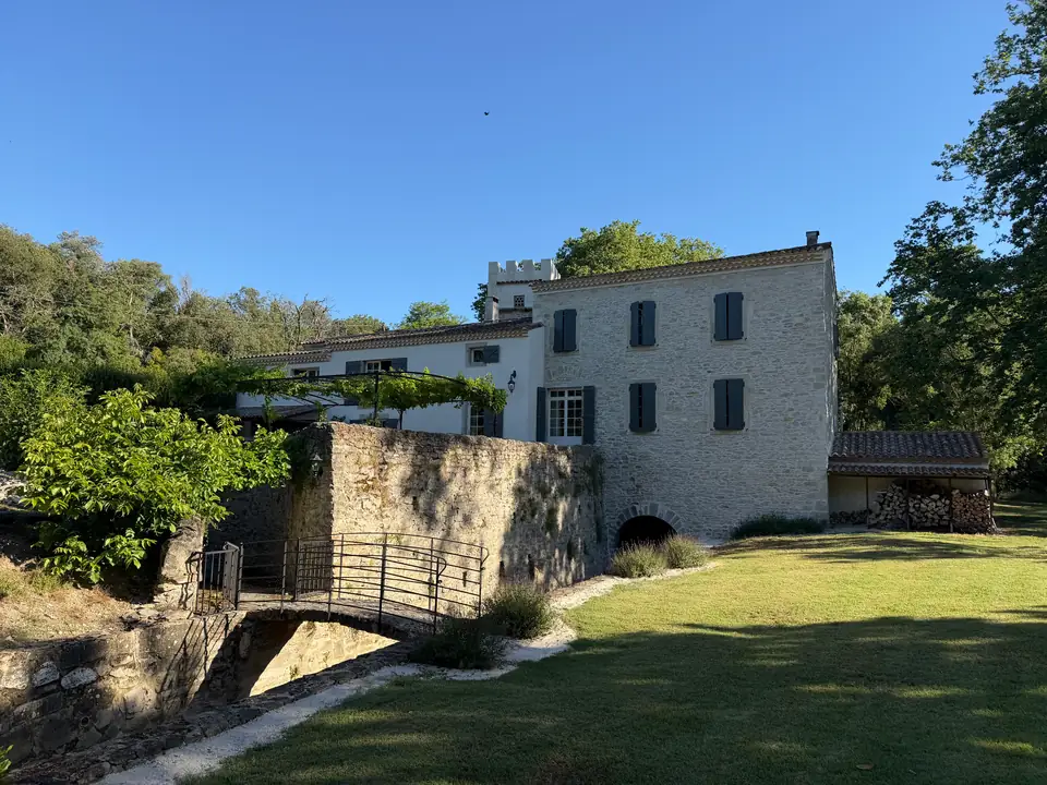 The back of the mill: a high stone retaining wall with an iron footbridge, the pergola above it and an arch at its foot.