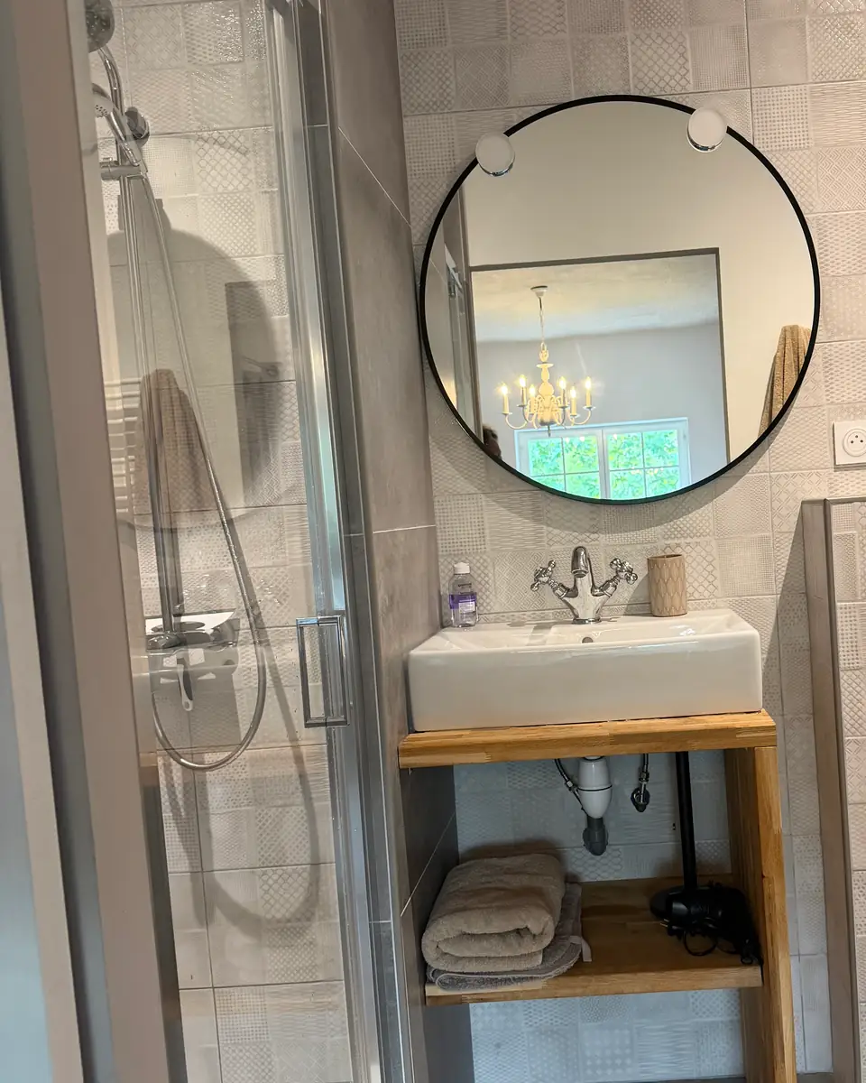 A bathroom with patterned grey tiles, a glass shower and a round black-framed mirror.