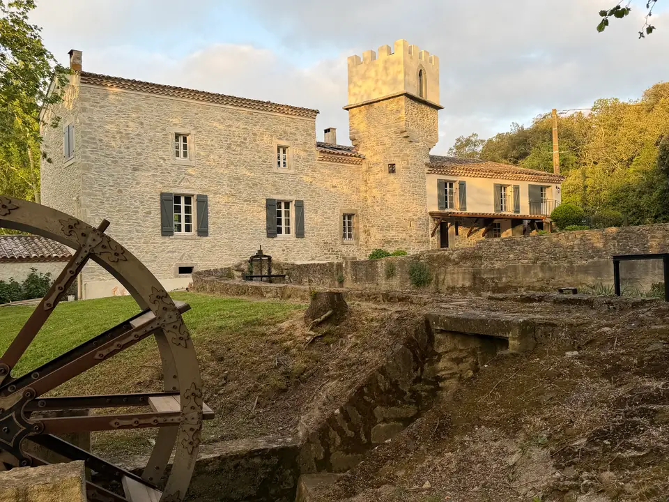 Evening sun on the long stone façade and the crenellated tower; on the left an iron wheel above a dry stone channel.