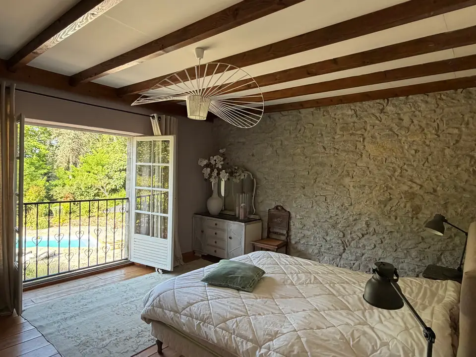 A bedroom with a stone wall and beams; French doors open onto an iron balcony above the pool.