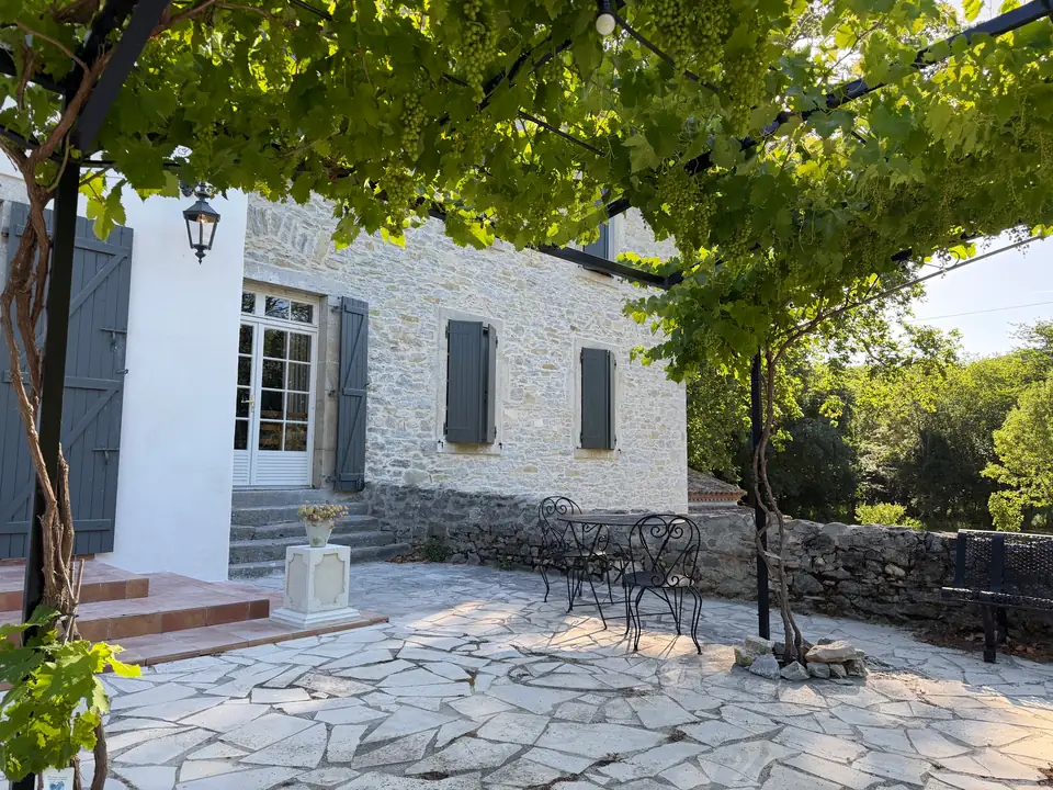 Grapes hang from the pergola; behind them the white wing and the stone house.