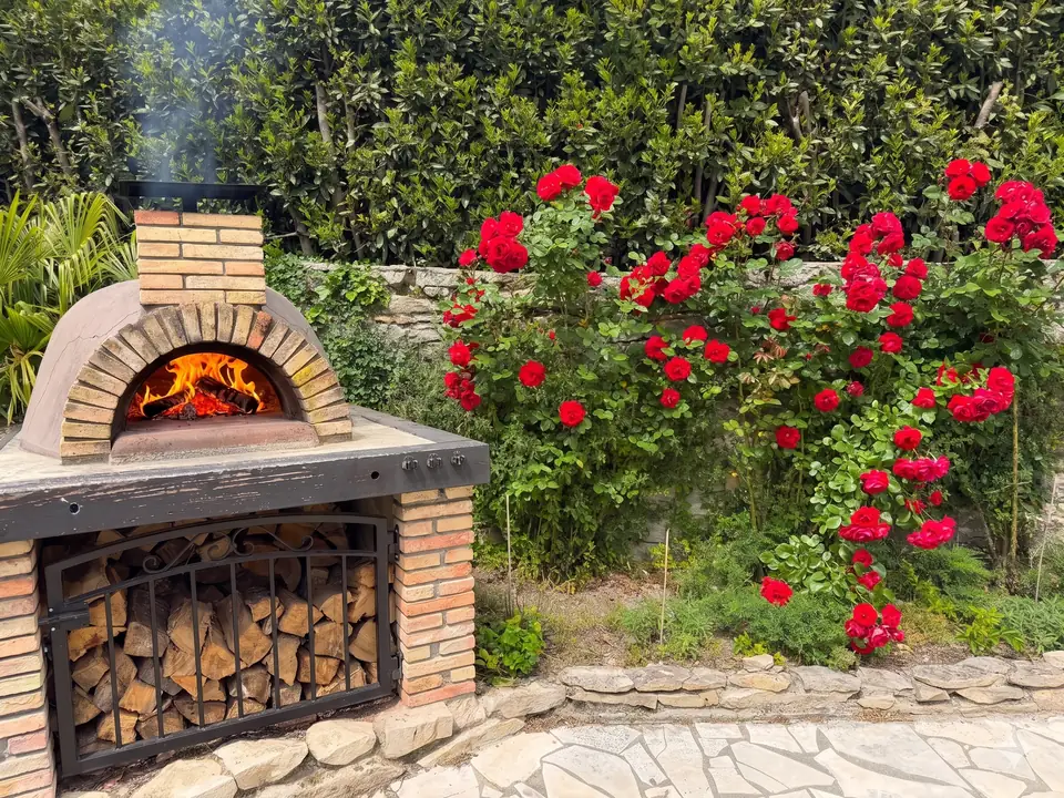 The pizza oven alight beside red climbing roses; the wood is stacked below behind an iron grille.