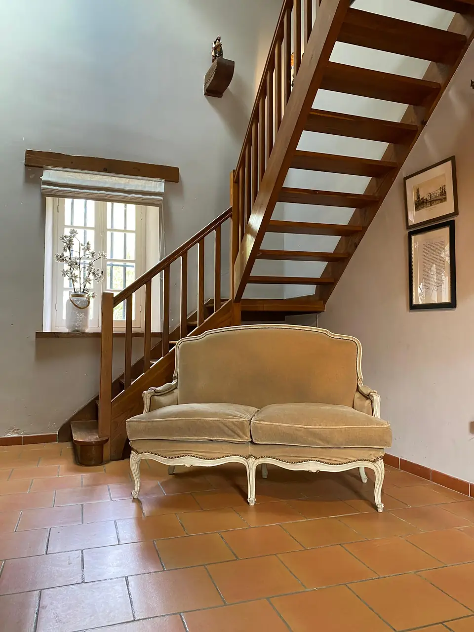 A velvet settee below the wooden staircase, beside a deep-set window with a blind.