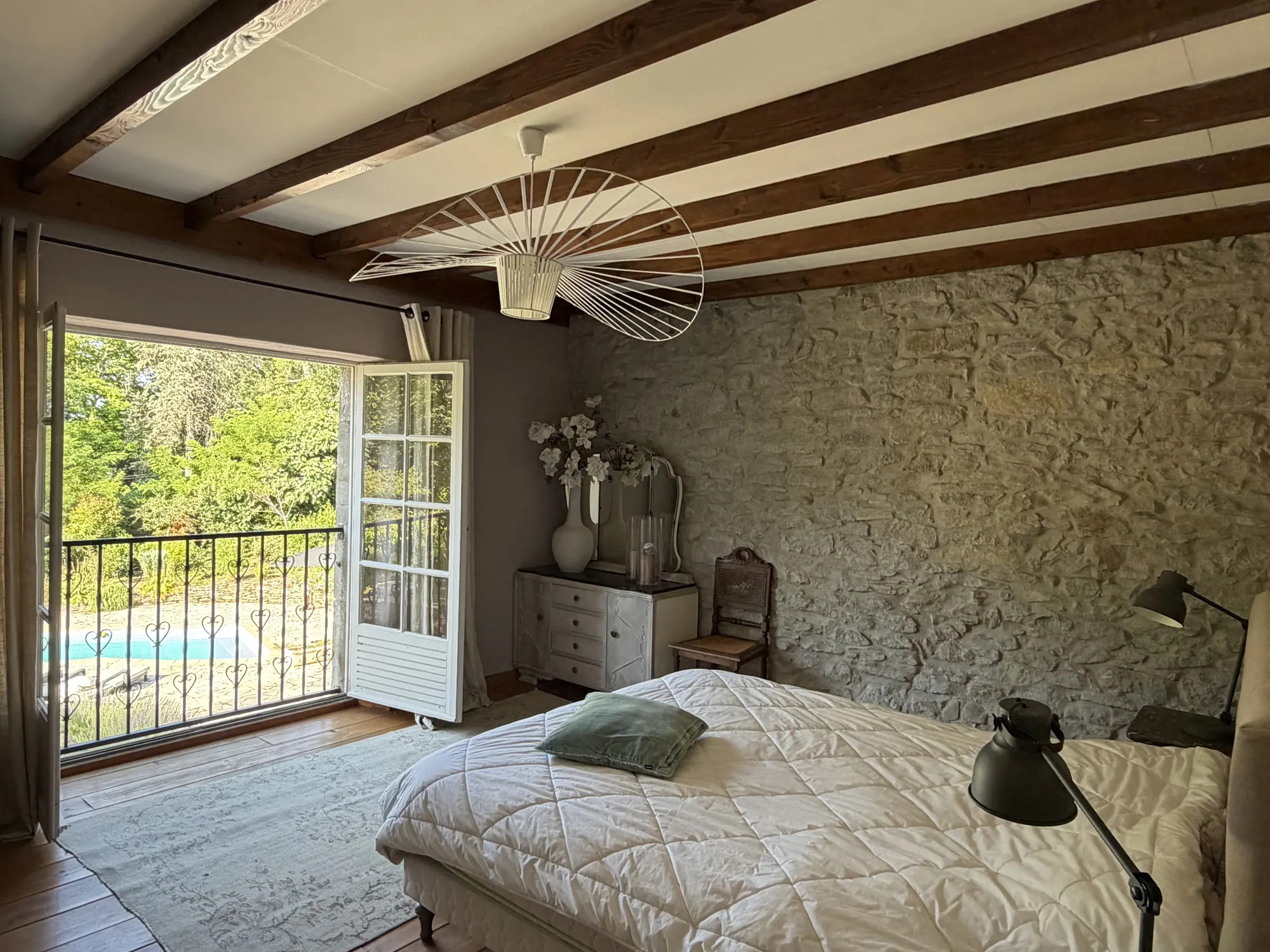 Een slaapkamer met balken en een stenen wand, met deuren naar een balkon boven het zwembad.